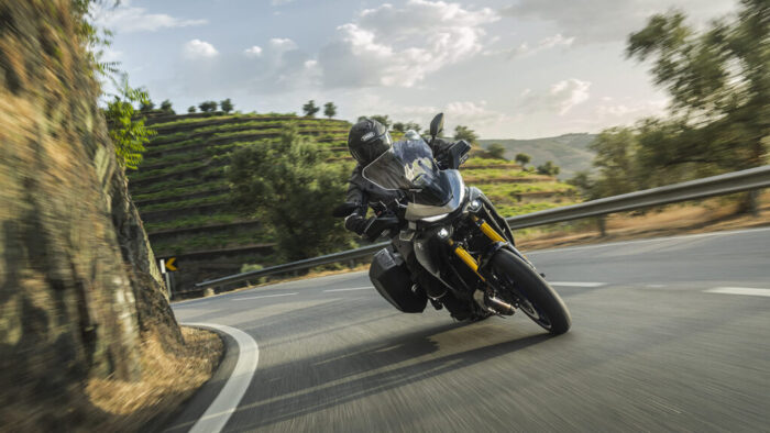 Yamaha Tracer 7 GT Y-AMT 2026 v globokem nagibu skozi ovinek, napredno vzmetenje, aerodinamična zaščita in potovalni kovčki v akciji.