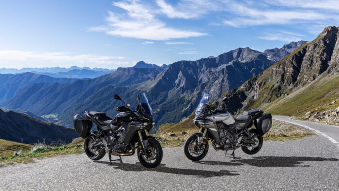 Dva motocikla Yamaha Tracer 9 GT v različnih barvah na razgledni gorski cesti z dramatično panoramo v ozadju.