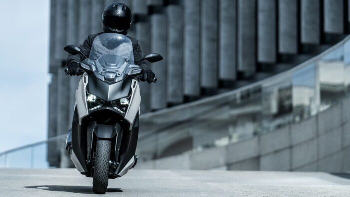 Sprednji pogled na Yamaha XMAX 300 Tech MAX+ med urbano vožnjo – samozavesten nastop, visoko nameščeni smerniki in zaščitni vizir za udobje na vsakem kilometru.