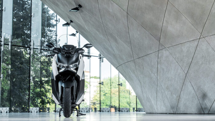 Yamaha XMAX 300 Tech MAX+ postavljen v sodobnem arhitekturnem ambientu – agresiven sprednji del z dvojno LED osvetlitvijo in prepoznavnim X-podpisom.